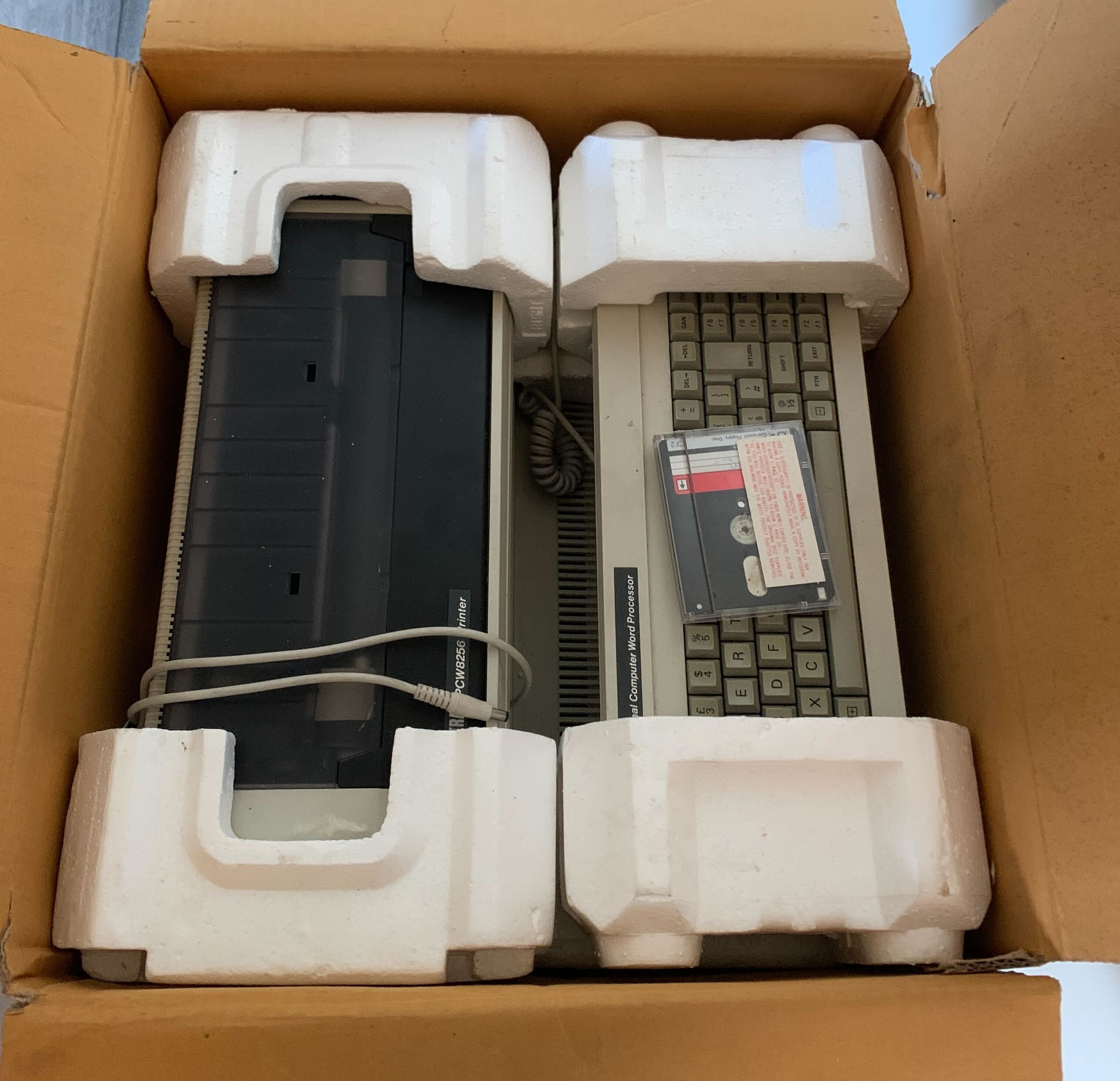 A view of an open cardboard box from above, showing a dot-matrix printer and keyboard in polystyrene packaging.
